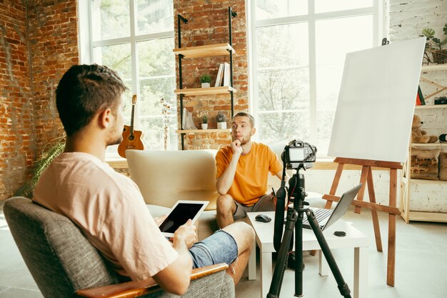 Dos jóvenes bloggers varones caucásicos en ropa casual con equipo profesional o cámara grabando video entrevista en casa. Blogs, videoblogs, vlogs. Hablar mientras se transmite en vivo en interiores.