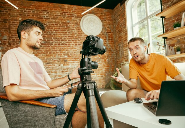 Dos jóvenes bloggers varones caucásicos en ropa casual con equipo profesional o cámara grabando video entrevista en casa. Blogs, videoblogs, vlogs. Hablar mientras se transmite en vivo en interiores.