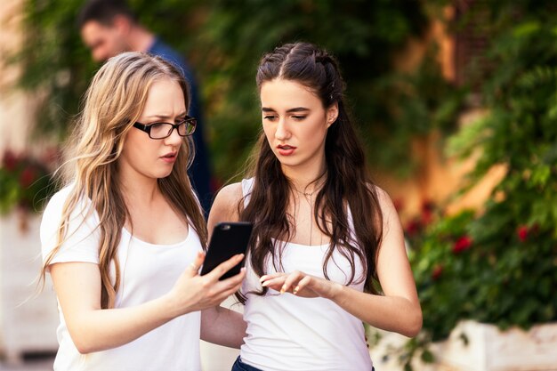 Dos jóvenes caucásicas están discutiendo algo y mirando en el teléfono inteligente con caras serias