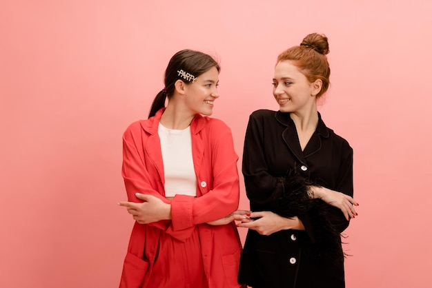 Foto gratuita dos jóvenes caucásicas positivas en trajes con las manos cruzadas se miran sobre fondo rosa tendencias de moda de primavera de estilo elegante