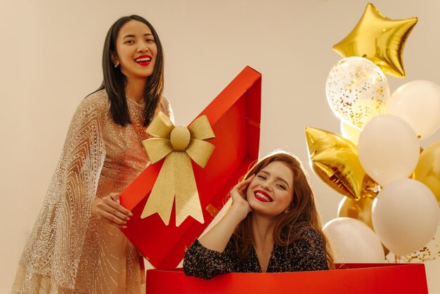 Dos jóvenes chicas interraciales, pelirroja y morena, sonríen a la cámara contra el fondo de globos y una gran caja roja de regalo.