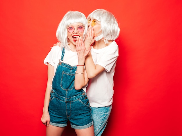 Dos jóvenes chicas sexy hipster con pelucas blancas y labios rojos. Hermosas mujeres de moda en ropa de verano. Modelos despreocupadas posando junto a la pared roja en verano de estudio comparte secretos, chismes