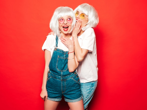 Dos jóvenes chicas sexy hipster con pelucas blancas y labios rojos. Hermosas mujeres de moda en ropa de verano. Modelos despreocupadas posando junto a la pared roja en verano de estudio comparte secretos, chismes