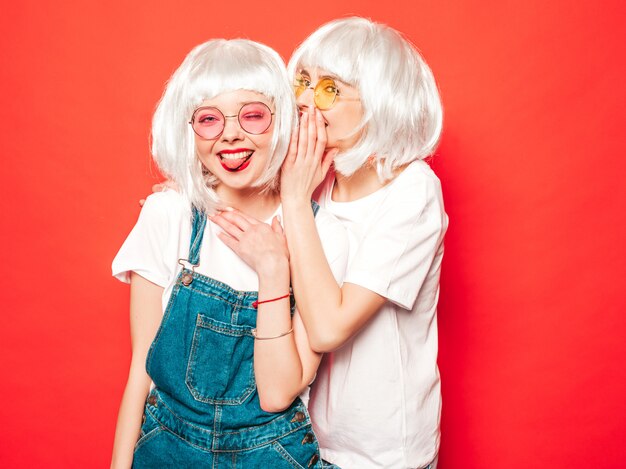 Dos jóvenes chicas sexy hipster con pelucas blancas y labios rojos. Hermosas mujeres de moda en ropa de verano. Modelos despreocupadas posando junto a la pared roja en verano de estudio comparte secretos, chismes