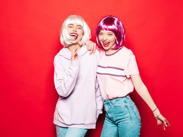 Dos jóvenes chicas sexy hipster sonrientes con pelucas blancas y labios rojos. Hermosas mujeres de moda en ropa de verano. Modelos despreocupados posando junto a la pared roja en el estudio volviéndose loco