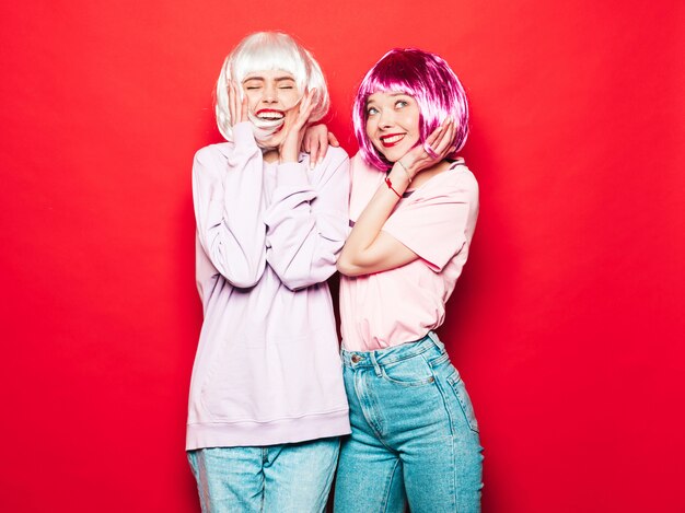 Dos jóvenes chicas sexy hipster sonrientes con pelucas blancas y labios rojos. Hermosas mujeres de moda en ropa de verano. Modelos despreocupados posando junto a la pared roja en el estudio volviéndose loco