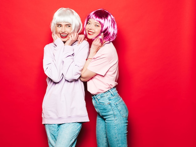 Dos jóvenes chicas sexy hipster sonrientes con pelucas blancas y labios rojos. Hermosas mujeres de moda en ropa de verano. Modelos despreocupados posando junto a la pared roja en el estudio volviéndose loco