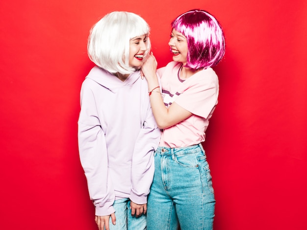 Dos jóvenes chicas sexy hipster sonrientes con pelucas blancas y labios rojos. Hermosas mujeres de moda en ropa de verano. Modelos despreocupados posando junto a la pared roja en el estudio volviéndose loco