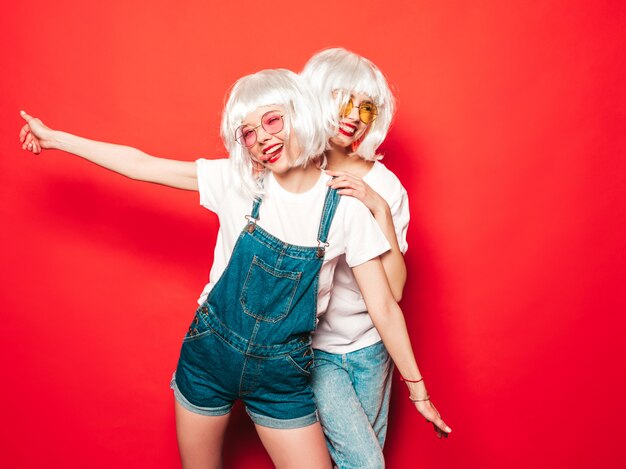 Dos jóvenes chicas sexy hipster sonrientes con pelucas blancas y labios rojos. Hermosas mujeres de moda en ropa de verano. Modelos despreocupados posando junto a la pared roja en verano de estudio con gafas de sol