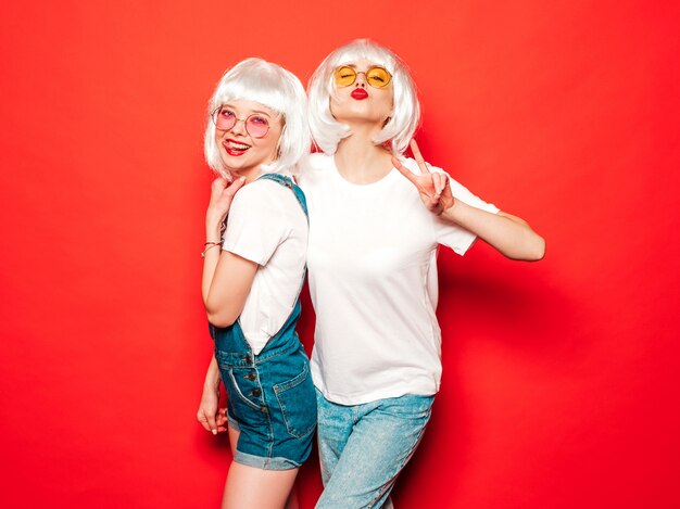 Dos jóvenes chicas sexy hipster sonrientes con pelucas blancas y labios rojos. Hermosas mujeres de moda en ropa de verano. Modelos despreocupados posando junto a la pared roja en verano de estudio volviéndose loco