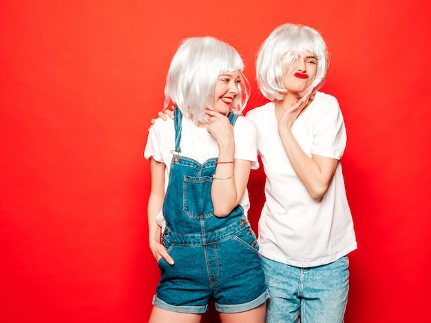 Dos jóvenes chicas sexy hipster sonrientes con pelucas blancas y labios rojos. Hermosas mujeres de moda en ropa de verano. Modelos despreocupados posando junto a la pared roja en verano de estudio volviéndose loco