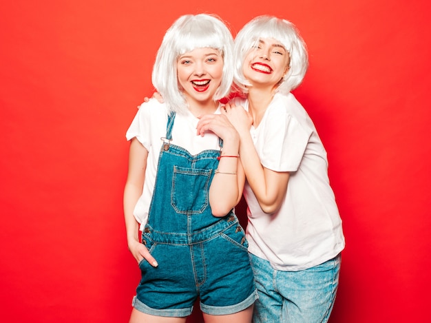 Dos jóvenes chicas sexy hipster sonrientes con pelucas blancas y labios rojos. Hermosas mujeres de moda en ropa de verano. Modelos despreocupados posando junto a la pared roja en verano de estudio volviéndose loco