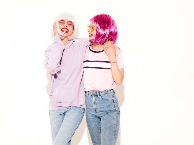 Dos jóvenes chicas sexy hipster sonrientes con pelucas y labios rojos. Hermosas mujeres de moda en ropa de verano. Modelos despreocupados posando junto a la pared blanca en estudio volviéndose locos y abrazándose