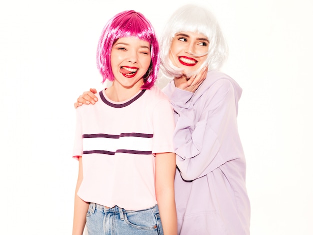 Dos jóvenes chicas sexy hipster sonrientes con pelucas y labios rojos. Hermosas mujeres de moda en ropa de verano. Modelos despreocupados posando junto a la pared blanca en estudio volviéndose locos y abrazándose