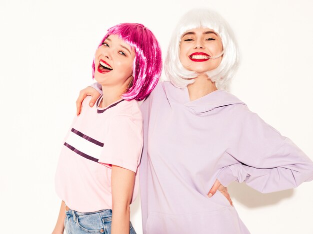 Dos jóvenes chicas sexy hipster sonrientes con pelucas y labios rojos. Hermosas mujeres de moda en ropa de verano. Modelos despreocupados posando junto a la pared blanca en estudio volviéndose locos y abrazándose