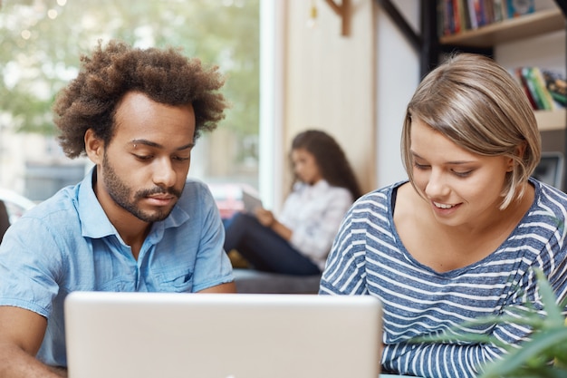 Dos jóvenes diseñadores independientes sentados en un espacio de trabajo conjunto, buscando en el monitor de una computadora portátil, buscando nuevas ideas para diseñadores de muebles. Par de jóvenes alegres sonriendo, gastando, mañana en libra