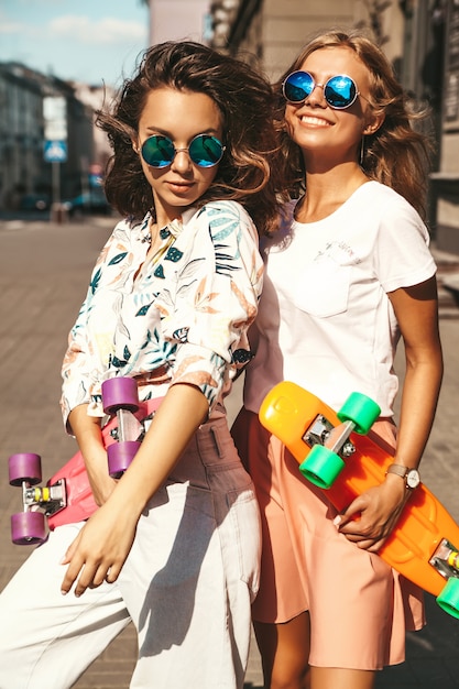 Dos jóvenes y elegantes modelos de mujeres rubias y hippies sonrientes en un día soleado de verano en ropa hipster con penny skateboard posando