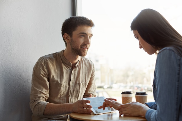 Dos jóvenes empresarios interesados en reunirse tomando café, hablando sobre un proyecto de puesta en marcha futura y mirando ejemplos de diseño de sitios web en tabletas en la cafetería. Productiva mañana en cómoda pla