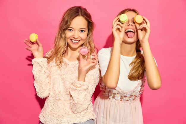 Dos jóvenes encantadoras hermosas mujeres hipster sonrientes en ropa de moda de verano. Mujeres haciendo gafas, gafas con coloridos macarons, sosteniendo macarons en el lugar de los ojos. Posando en pared rosa