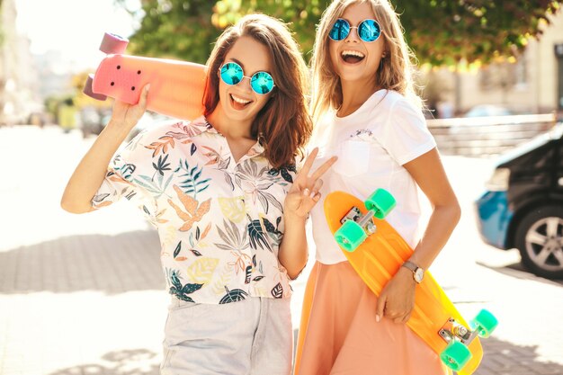 Dos jóvenes con estilo hippie sonriente morena y rubia mujeres modelos en ropa hipster de verano con penny skateboard posando. Cara sorpresa, emociones