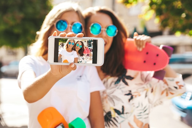 Dos jóvenes con estilo sonriente hippie morena en día soleado de verano en ropa hipster con penny skateboard posando