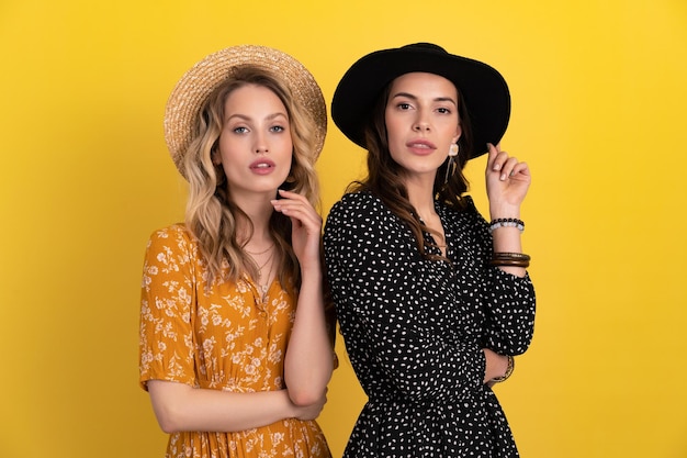 Dos jóvenes hermosas amigas juntas aisladas sobre fondo amarillo en vestido negro y amarillo y sombrero con estilo boho tendencia primavera verano accesorios de estilo de moda confiados sexy