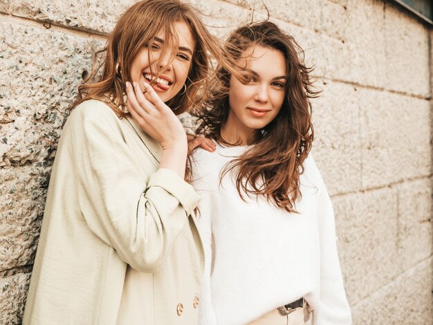 Dos jóvenes hermosas chicas hipster sonrientes en abrigo y suéter blanco de moda