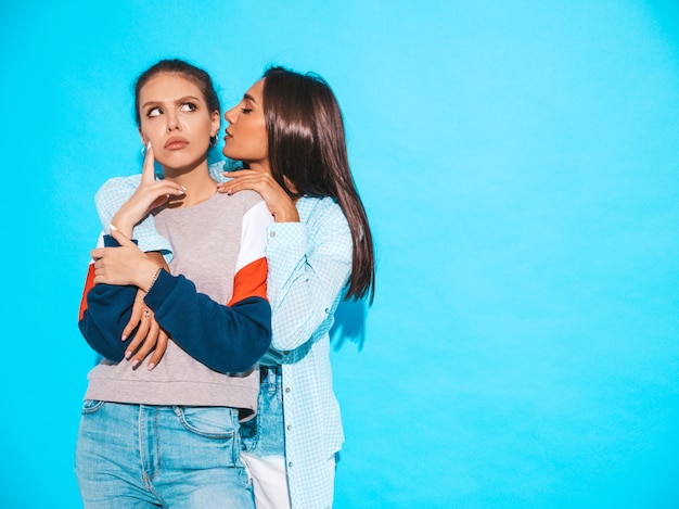 Dos jóvenes hermosas chicas hipster sonrientes en ropa casual de moda de verano. Las mujeres sexy comparten secretos, chismes. Aisladas en azul. Emociones faciales sorprendidas