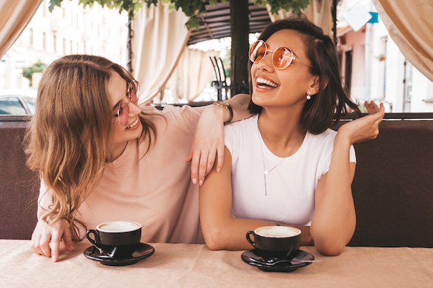 Dos jóvenes hermosas chicas hipster sonrientes en ropa casual de verano de moda. Mujeres despreocupadas charlando en la terraza terraza café y tomando café. Modelos positivos divirtiéndose y comunicándose
