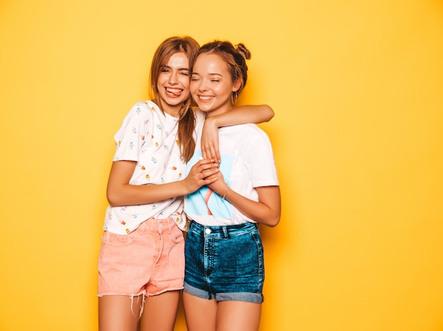 Dos jóvenes hermosas chicas hipster sonrientes en ropa de moda de verano. Mujeres despreocupadas atractivas que presentan cerca de la pared amarilla. Modelos positivos volviéndose locos y divirtiéndose.