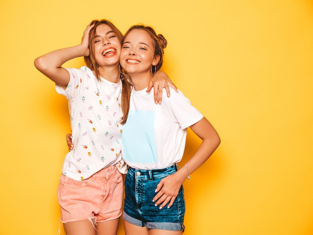 Dos jóvenes hermosas chicas hipster sonrientes en ropa de moda de verano. Mujeres despreocupadas atractivas que presentan cerca de la pared amarilla. Modelos positivos volviéndose locos y divirtiéndose.