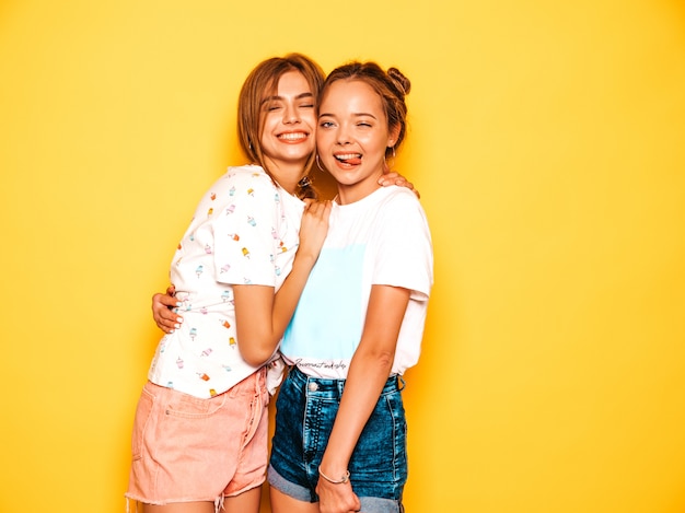 Dos jóvenes hermosas chicas hipster sonrientes en ropa de moda de verano. Mujeres despreocupadas atractivas que presentan cerca de la pared amarilla. Modelos positivos volviéndose locos y divirtiéndose.