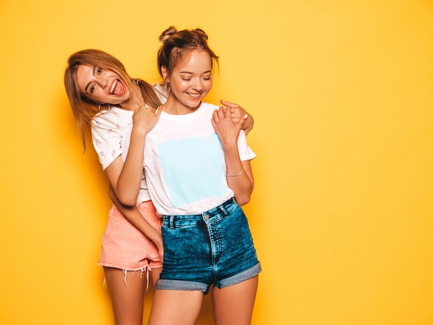 Dos jóvenes hermosas chicas hipster sonrientes en ropa de moda de verano. Mujeres despreocupadas atractivas que presentan cerca de la pared amarilla. Modelos positivos volviéndose locos y divirtiéndose.