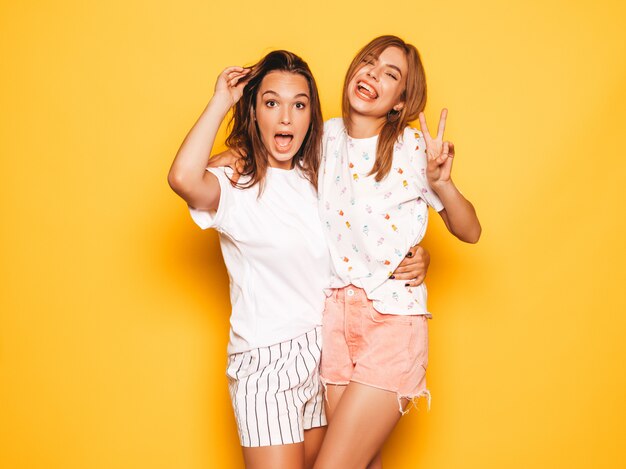 Dos jóvenes hermosas chicas hipster sonrientes en ropa de moda de verano. Mujeres despreocupadas atractivas que presentan cerca de la pared amarilla. Modelos positivos volviéndose locos y divirtiéndose.