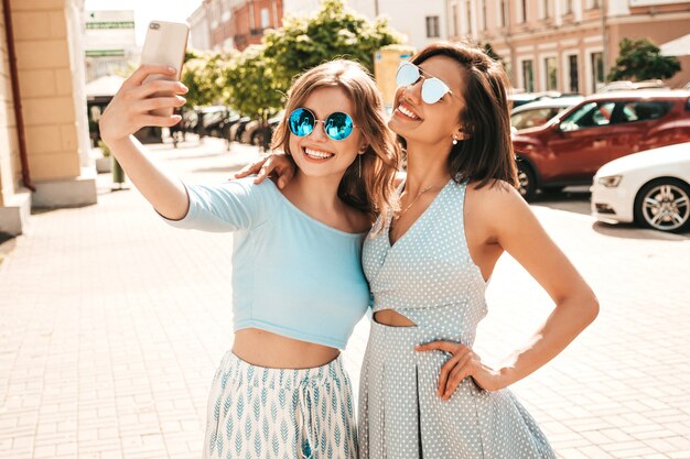 Dos jóvenes hermosas chicas hipster sonrientes en ropa de moda de verano. Mujeres despreocupadas sexy posando en el fondo de la calle en gafas de sol. Ellos toman fotos de autorretratos en el teléfono inteligente al atardecer