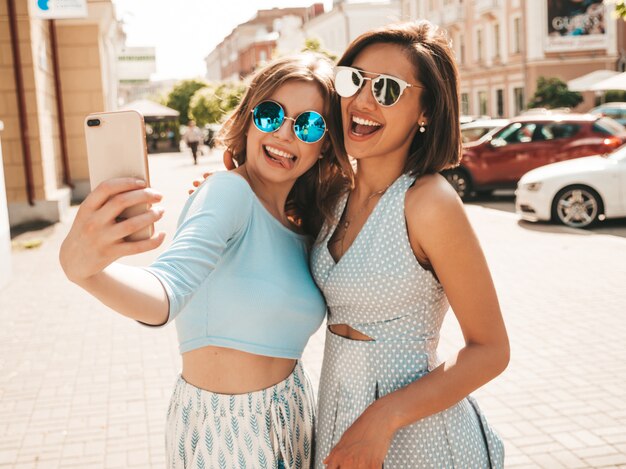 Dos jóvenes hermosas chicas hipster sonrientes en ropa de moda de verano. Mujeres despreocupadas sexy posando en el fondo de la calle en gafas de sol. Ellos toman fotos de autorretratos en el teléfono inteligente al atardecer