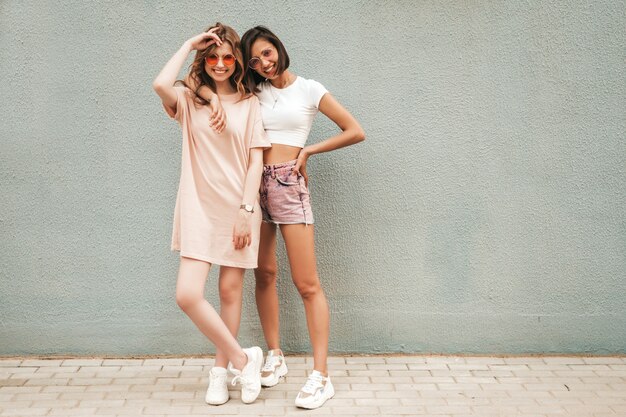 Dos jóvenes hermosas chicas hipster sonrientes en ropa de moda de verano. Mujeres despreocupadas sexy posando en el fondo de la calle en gafas de sol. Modelos positivos divirtiéndose y abrazándose