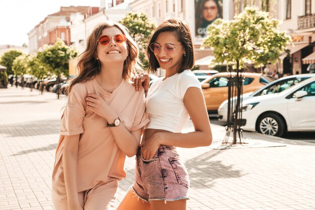 Dos jóvenes hermosas chicas hipster sonrientes en ropa de moda de verano. Mujeres despreocupadas sexy posando en el fondo de la calle en gafas de sol. Modelos positivos divirtiéndose y volviéndose locos