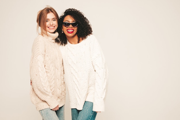 Dos jóvenes hermosas chicas hipster sonrientes en suéteres de invierno de moda. Modelos positivos divirtiéndose y abrazándose.