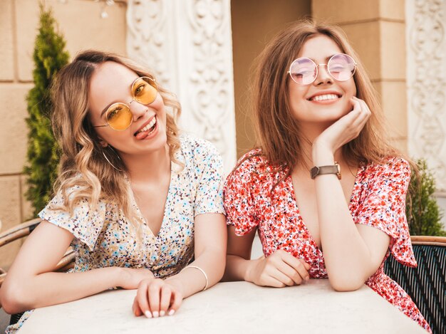 Dos jóvenes hermosas chicas hipster sonrientes en vestido de verano de moda. Mujeres despreocupadas charlando en el café de la terraza en el fondo de la calle con gafas de sol. Modelos positivos divirtiéndose y comunicándose