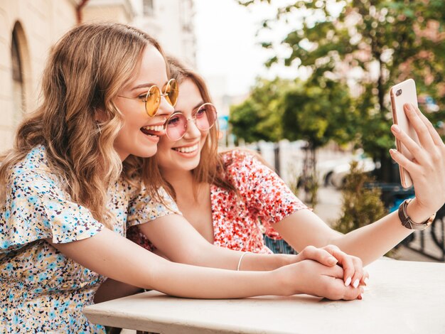 Dos jóvenes hermosas chicas hipster sonrientes en vestido de verano de moda. Mujeres despreocupadas charlando en el café de la terraza en el fondo de la calle. Modelos positivos divirtiéndose y tomando selfie en teléfono inteligente