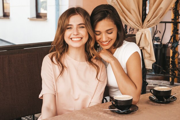 Dos jóvenes hermosas chicas hipster sonrientes en vestido de verano de moda. Mujeres despreocupadas charlando en el café de la terraza y tomando café. Modelos positivos divirtiéndose y comunicándose