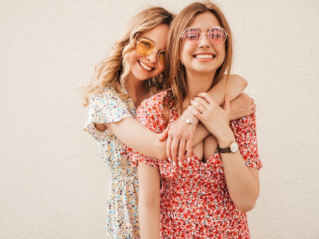 Dos jóvenes hermosas chicas hipster sonrientes en vestido de verano de moda. Mujeres despreocupadas sexy posando en la calle cerca de la pared en gafas de sol. Modelos positivos divirtiéndose y volviéndose locos