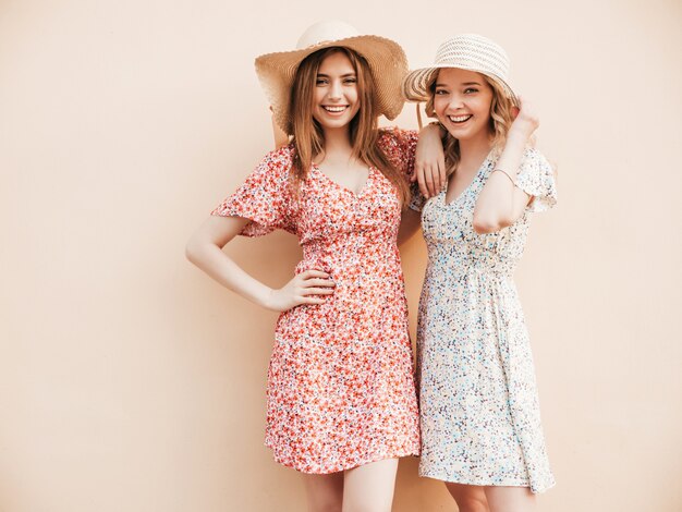 Dos jóvenes hermosas chicas hipster sonrientes en vestido de verano de moda. Mujeres despreocupadas sexy posando en la calle junto a la pared con sombreros. Modelos positivos divirtiéndose y abrazándose