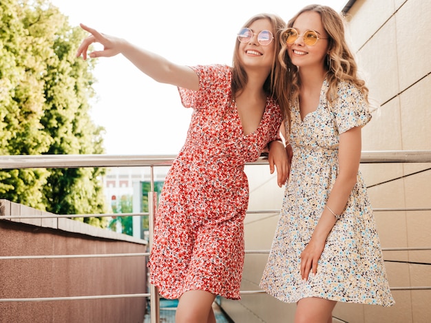 Dos jóvenes hermosas chicas hipster sonrientes en vestido de verano de moda. Mujeres despreocupadas sexy posando en el fondo de la calle en gafas de sol. Modelos positivos divirtiéndose y señalando algo interesante