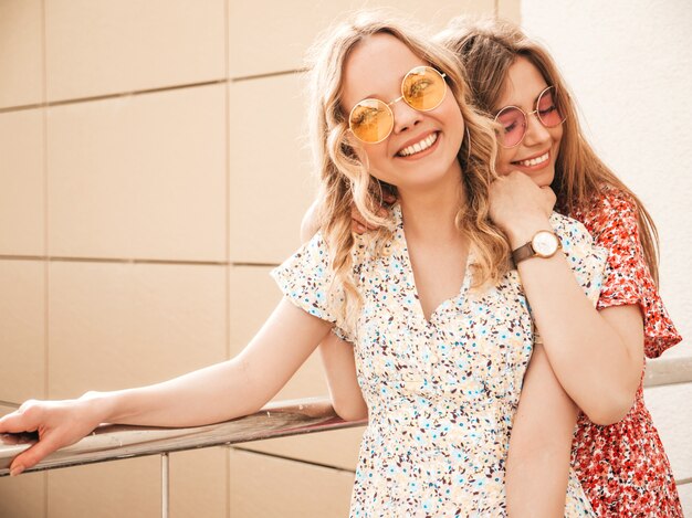 Dos jóvenes hermosas chicas hipster sonrientes en vestido de verano de moda. Mujeres despreocupadas sexy posando en el fondo de la calle en gafas de sol. Modelos positivos divirtiéndose y volviéndose locos