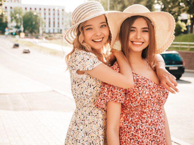 Dos jóvenes hermosas chicas hipster sonrientes en vestido de verano de moda. Mujeres despreocupadas sexy posando en el fondo de la calle con sombreros. Modelos positivos divirtiéndose y abrazándose. Se están volviendo locos