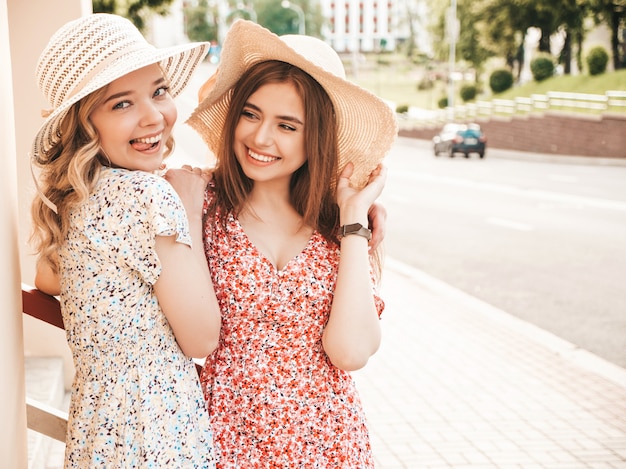 Dos jóvenes hermosas chicas hipster sonrientes en vestido de verano de moda. Mujeres despreocupadas sexy posando en el fondo de la calle con sombreros. Modelos positivos divirtiéndose y abrazándose