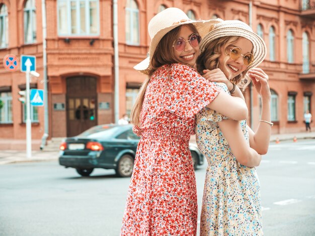 Dos jóvenes hermosas chicas hipster sonrientes en vestido de verano de moda. Mujeres despreocupadas sexy posando en el fondo de la calle con sombreros. Modelos positivos divirtiéndose y abrazándose