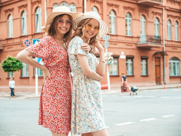 Dos jóvenes hermosas chicas hipster sonrientes en vestido de verano de moda. Mujeres despreocupadas sexy posando en el fondo de la calle con sombreros. Modelos positivos divirtiéndose y abrazándose